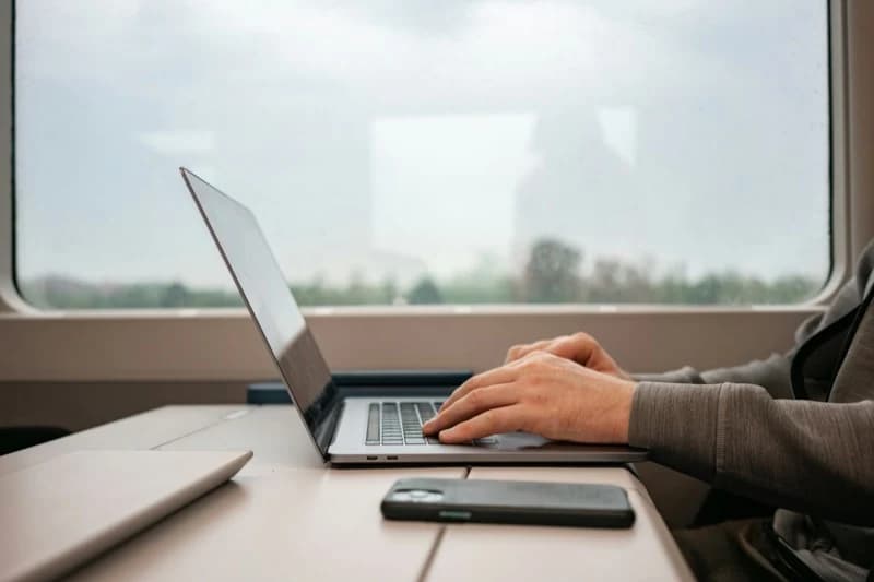 Hands typing on a silver laptop on a train table, with a phone resting beside it and an overcast morning landscape passing outside the window.